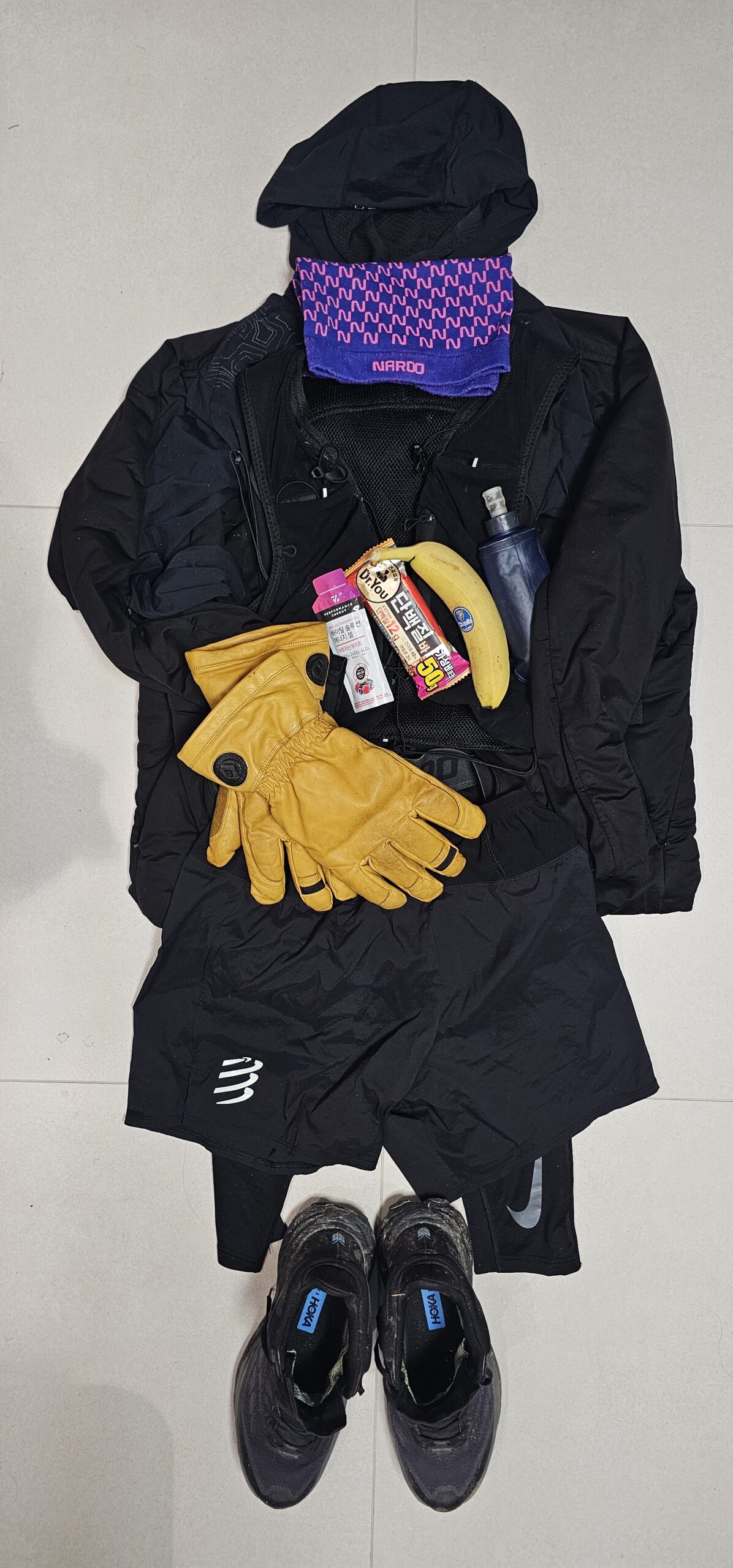 A flat-lay layout of winter trail running gear including gloves, neck warmer, hydration bottle, energy bar, banana, jacket, shorts, and trail running shoes.