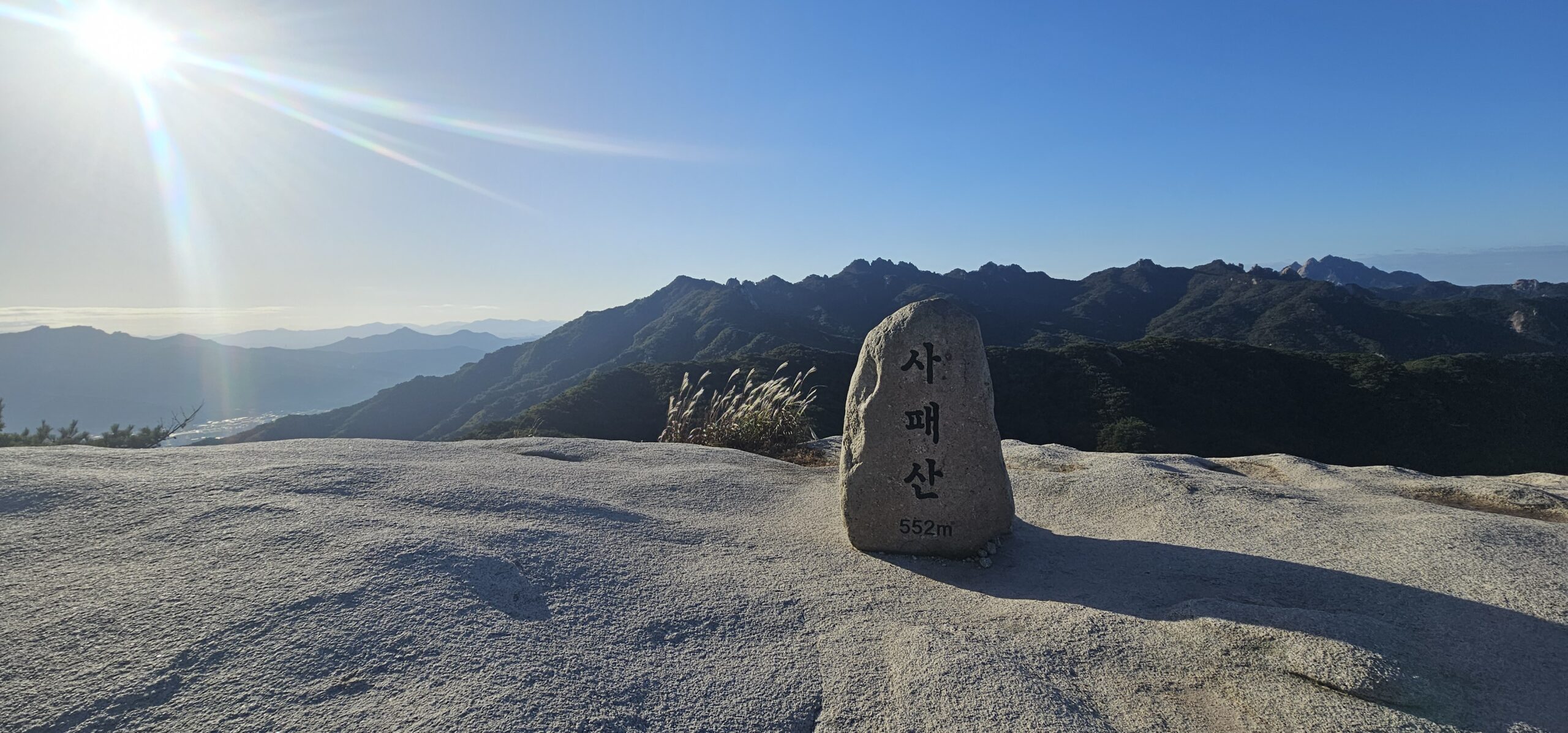 A stone summit marker engraved with “사패산 552m” standing on a granite surface, with a panoramic view of mountain ridges under clear blue sky.